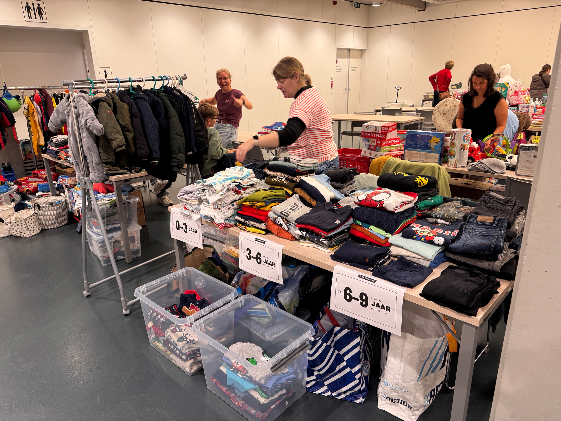 Mensen bij kledingtafels met leeftijdslabels en rekken in een zaal.
